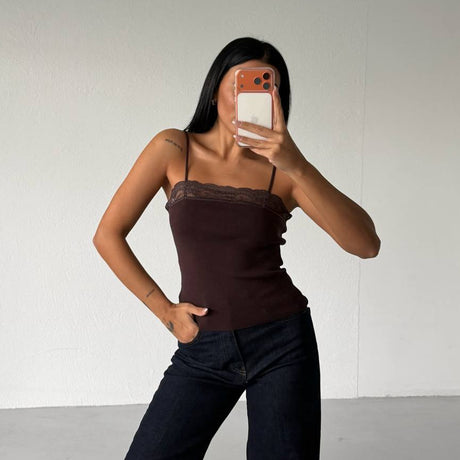 Person wearing a brown top and dark jeans taking a mirror selfie against a plain white wall.