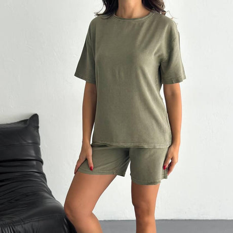 Person wearing a green t-shirt and shorts set with white sneakers in a minimalistic setting.