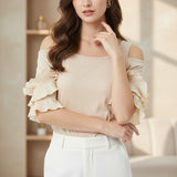 Woman wearing a beige off-shoulder top and white pants in a bright room.