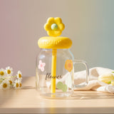 Clear mug with yellow flower lid and straw on a wooden surface with flowers and a cloth.
