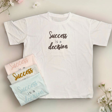 White t-shirt with 'Success is a decision' text, surrounded by smaller versions of the shirt and flowers on a light background.