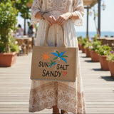Jute bag with 'Sun Salt Sandy' text and palm tree design held in front of a display of handbags.