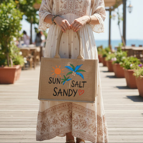 Jute bag with 'Sun Salt Sandy' text and palm tree design held in front of a display of handbags.