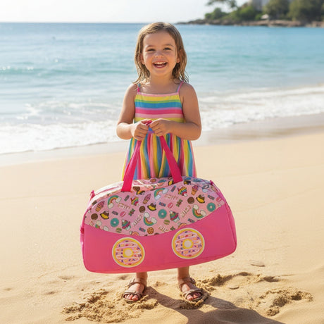 Young girl in a colorful dress holding a pink beach bag with fruit pattern on a sandy beach.