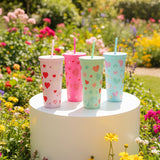 Four colorful tumblers with heart patterns on a white pedestal in a garden setting.