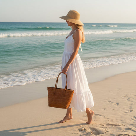 Woman in a white dress and hat walking on a beach with a woven bag.