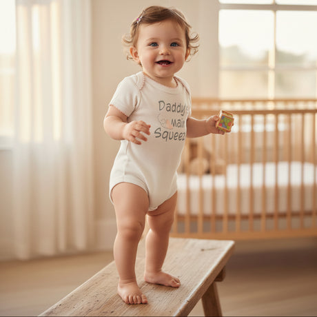 A baby standing on a wooden bench in a softly lit nursery, wearing a white short-sleeve bodysuit with the text 'Daddy's Mail Squeeez' and holding a small colorful toy.