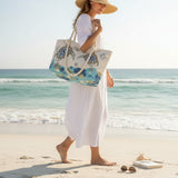 Woman on a beach holding a tote bag with turtle design, wearing a white dress and straw hat.