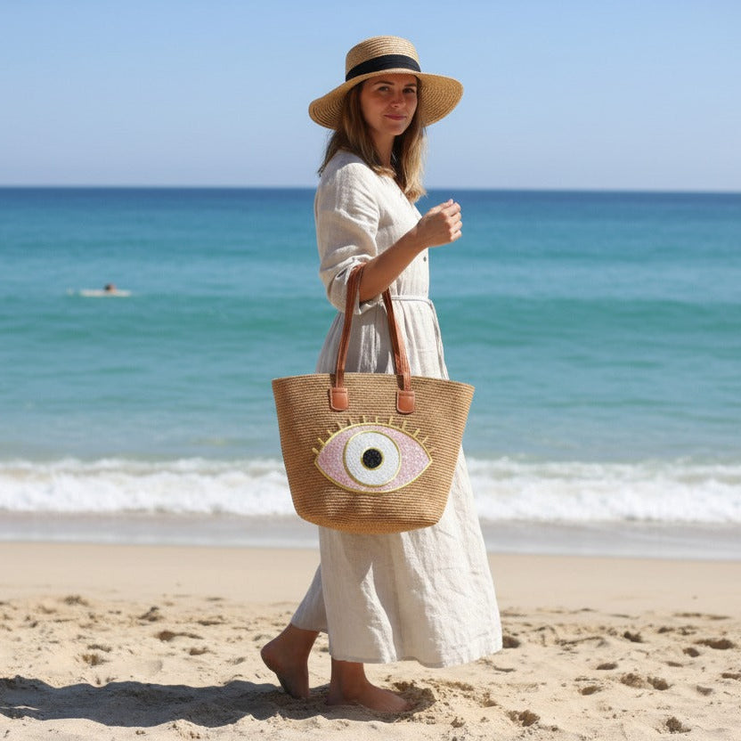 Woman on a beach holding a woven bag with an eye design, wearing a wide-brimmed hat.