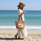 Woman on a beach holding a woven bag with an eye design, wearing a wide-brimmed hat.