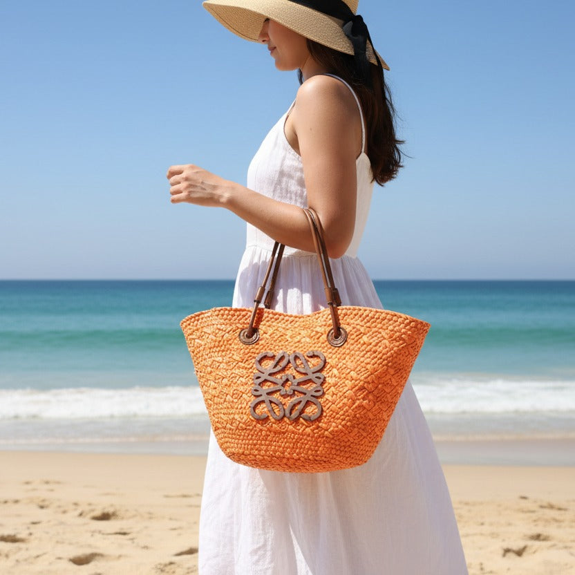 Woman holding an orange straw bag on a beach with blue sky and ocean.