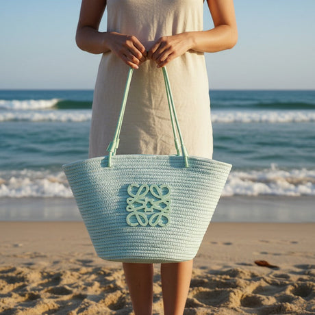 Person holding a light blue beach bag on a sandy beach with ocean waves in the background