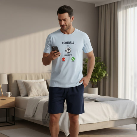 Man in a bedroom wearing a light blue t-shirt with text and graphics, holding a phone.
