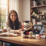 Woman sitting at a desk with a teddy bear in a graduation outfit and a laptop showing a video call.