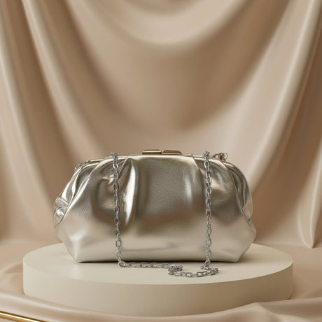 A metallic silver clutch bag with a gold chain strap, displayed on a round white pedestal against a draped beige satin background.