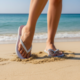 Person wearing white flip-flops on a sandy beach with ocean in the background