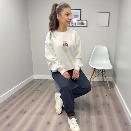 Woman wearing a white sweatshirt with a white bear design, dark pants, and white sneakers in a grey room