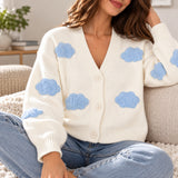 White knit cardigan with fuzzy blue cloud patches and a V neck, worn sitting with light jeans