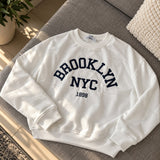 White sweatshirt with a navy Brooklyn NYC 1898 varsity print laid flat on a grey sofa