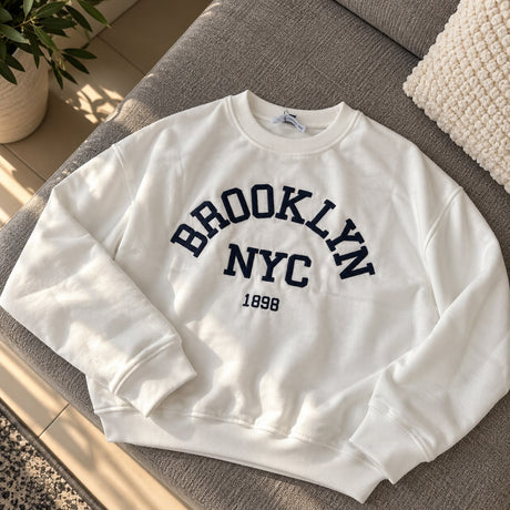 White sweatshirt with a navy Brooklyn NYC 1898 varsity print laid flat on a grey sofa