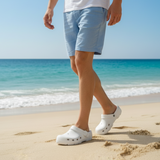 Person wearing light blue shorts and white clogs walking on a sandy beach with ocean in the background