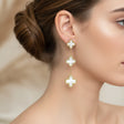 Gold and white clover-shaped earrings worn by a woman on a blurred background