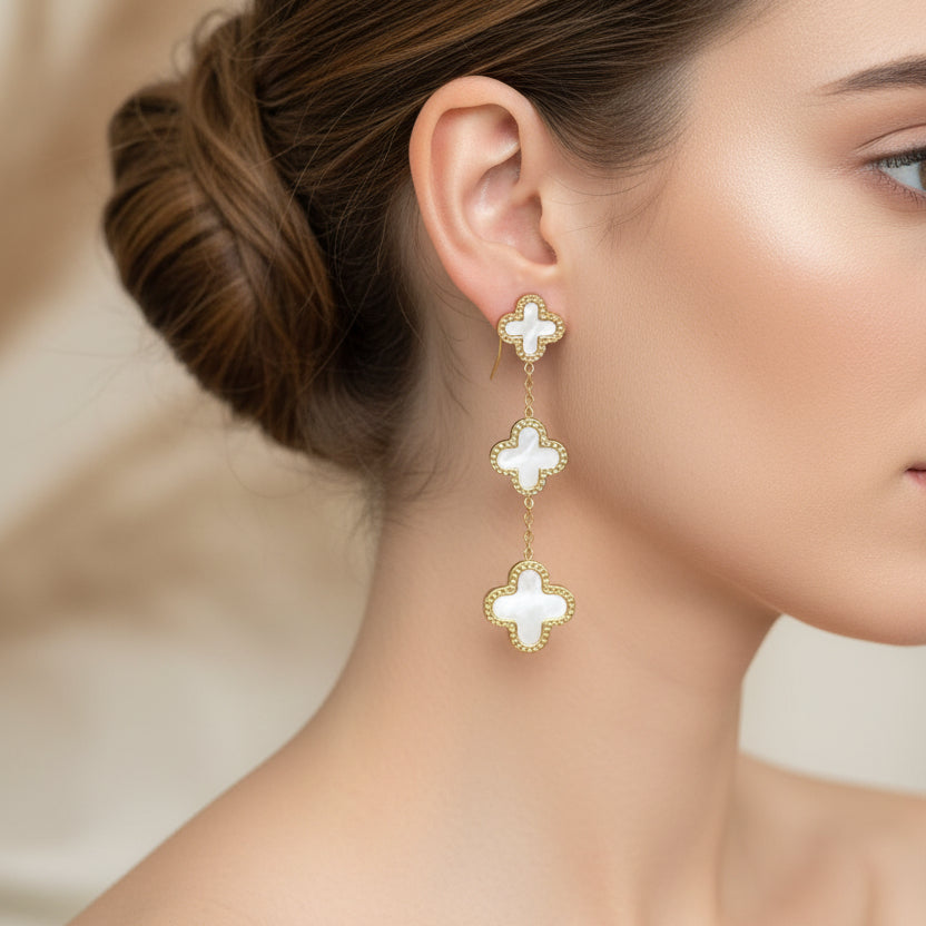 Gold and white clover-shaped earrings worn by a woman on a blurred background