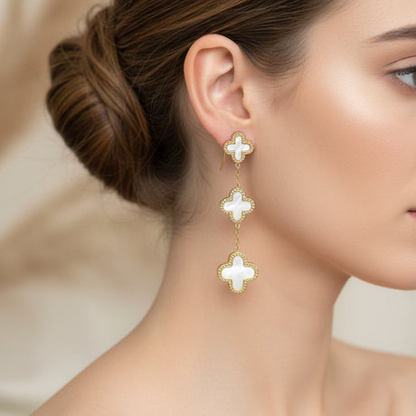 Gold and white clover-shaped earrings worn by a woman on a blurred background