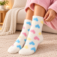 Fuzzy white kids socks covered in blue, pink and yellow hearts, worn on a cream rug