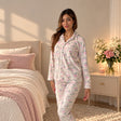 Woman in a white button up pyjama set with pink roses and grey leaves beside a pink floral bed
