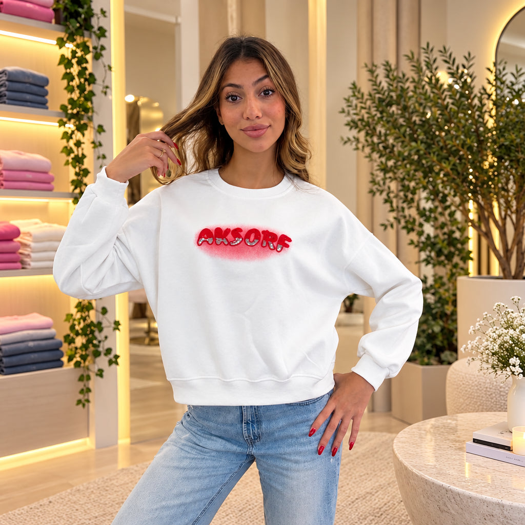 Woman in a white sweatshirt with red sequin lettering, hand in her hair, worn with light jeans