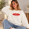 Woman sitting on a rug in a white sweatshirt with red and silver sequin letters