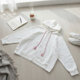 White zip hoodie laid flat on a pale wood floor with pink tassel drawstrings