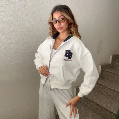 Woman in a white cropped zip hoodie with a navy letter patch and sunglasses on a staircase