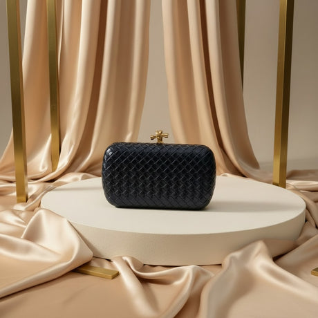 A navy woven clutch bag with a gold cross clasp sits on a round white pedestal