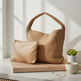 Woven bag and beige pouch on a wooden surface with a plant and cup in the background