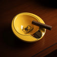 Deep round ash bowl in glossy yellow, a cigar laid across its gold lined notch