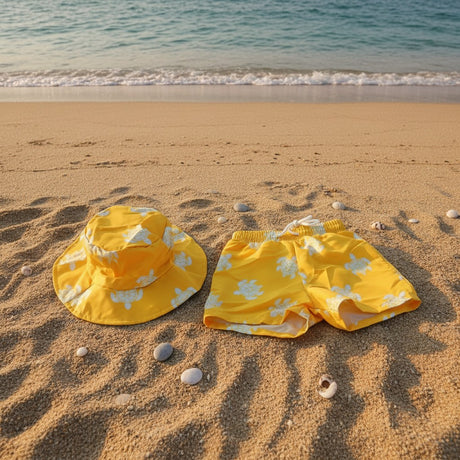 Yellow swim trunks and matching sun hat with a white floral pattern, laid out on a sandy beach near the shoreline