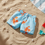 Light blue swim trunks with an orange lobster print, laid flat on a sandy beach with seashells and a striped towel nearby.