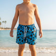 A child standing on a sandy beach wearing blue swim trunks with a black Mickey Mouse pattern and a white drawstring.