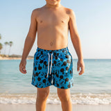 A child standing on a sandy beach wearing blue swim trunks with a black Mickey Mouse pattern and a white drawstring.