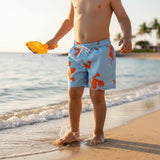 A child standing on a sandy beach at the water's edge wearing light blue swim trunks with an orange dinosaur pattern and holding a yellow shovel.