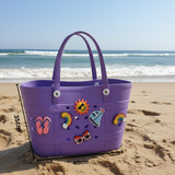 Purple beach bag with decorative stickers on a sandy beach with ocean waves in the background