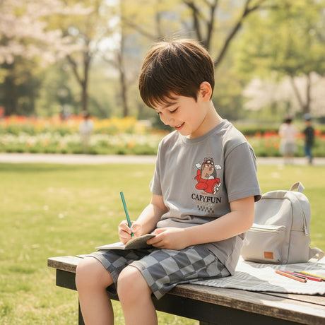 Child sitting on a bench in a park, writing in a notebook with a backpack next to them.