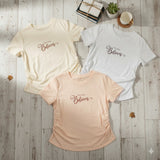 Three t-shirts in beige, white, and pink on a wooden surface with decorative items.
