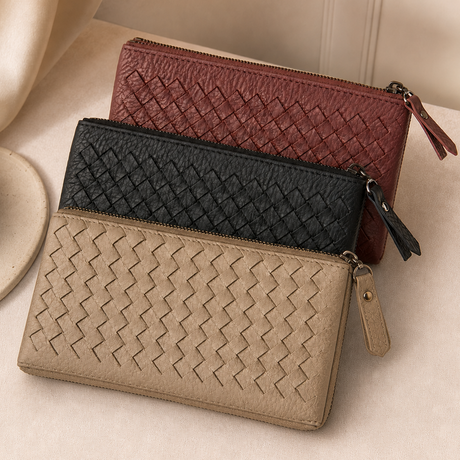 Three woven zip-around wallets in beige, black, and burgundy, arranged diagonally on a soft cream fabric surface.